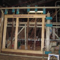 Timber door or window frame under construction with angled bracing supports, set within the workshop environment.